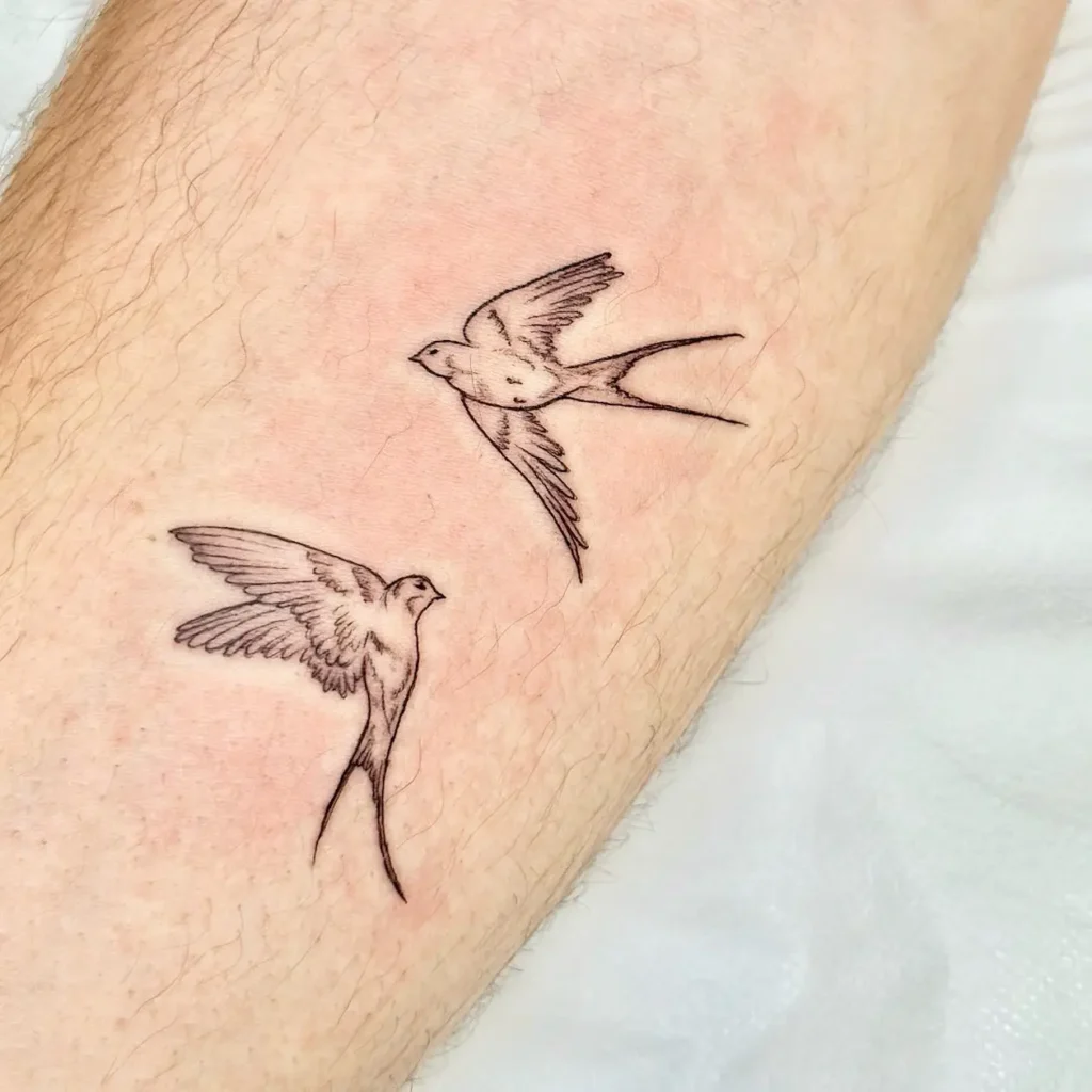 Fine line tattoo of two flying swallows with detailed wings and long tail feathers on light skin.