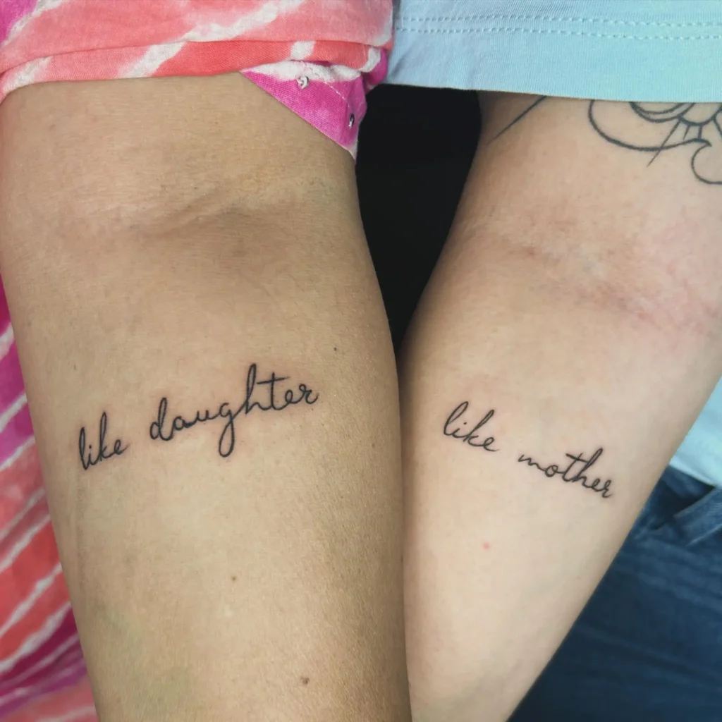 Two arms side by side, each with a cursive script tattoo. One arm reads “like daughter,” while the other says “like mother.” Both tattoos use clean, delicate black ink lines. This minimal design highlights the unbreakable link and shared identity found in mom and daughter tattoos, making it heartfelt and memorable.