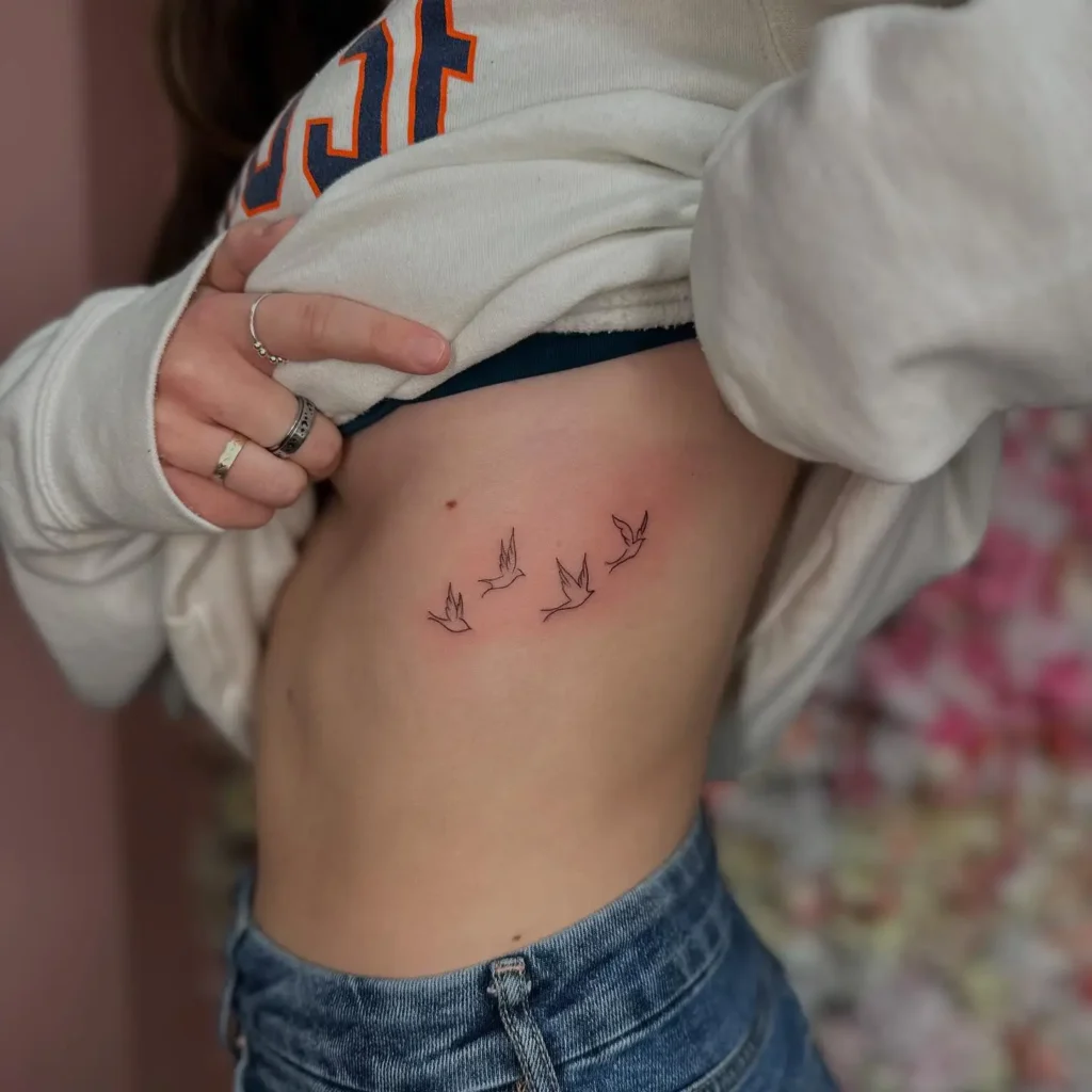 Minimalist tattoo of five flying birds in outline style on the side ribcage of a woman in jeans and sweatshirt.