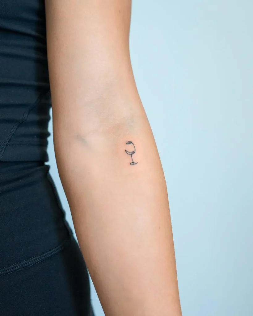Small black outline wine glass tattoo inked on the inside of a woman’s arm, near the elbow.