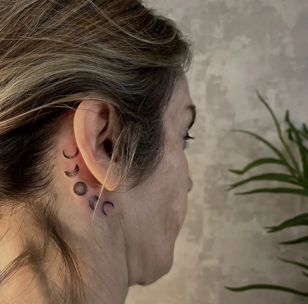 A vertical moon phase tattoo behind a woman’s ear, showing crescent, half, full, and waning moon shapes in black shading, positioned along the neck against light brown hair and neutral background.









