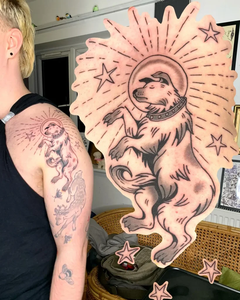 Tattoo of a dog in an astronaut helmet, floating among stars and cosmic rays on the upper arm, designed in a bold blackwork style for dog tattoo ideas.