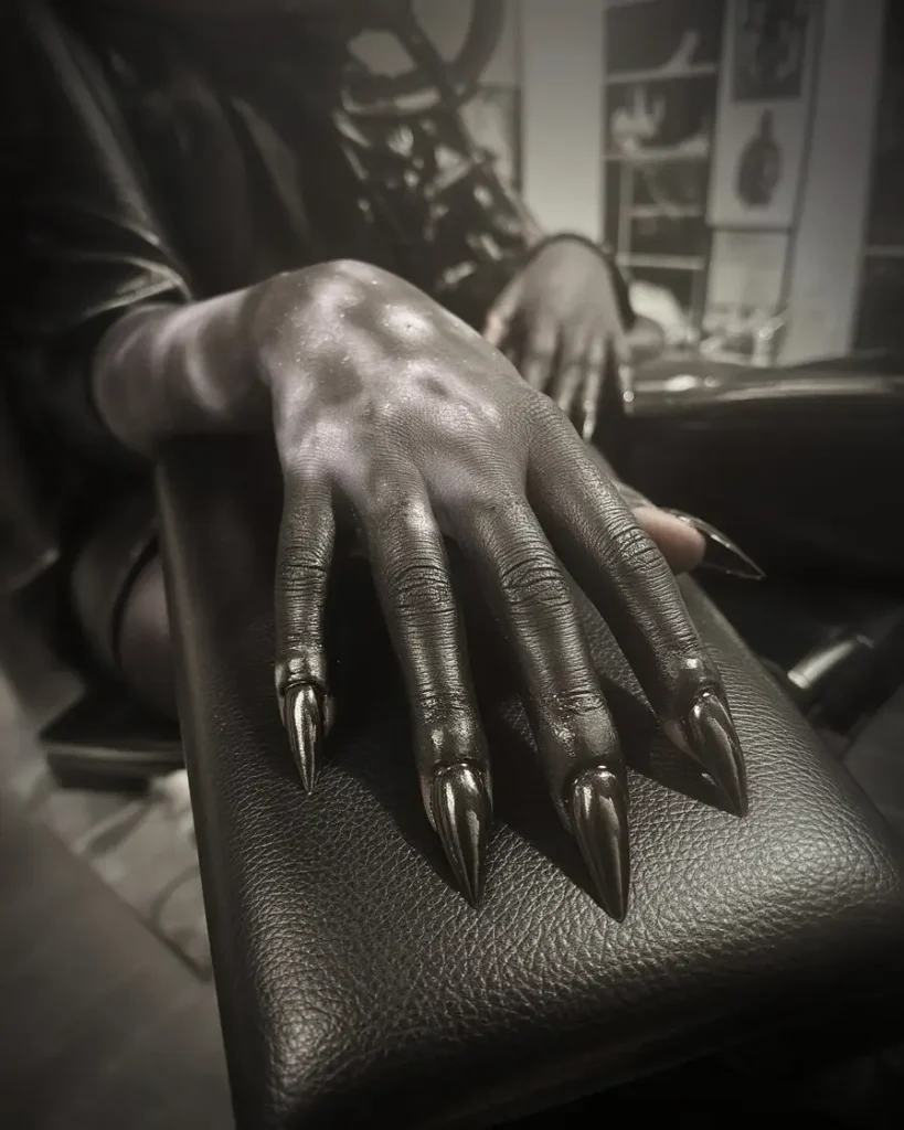 A hand with a gothic black tattoo and sharp claw-like nails, resting on a leather surface in a dimly lit setting.