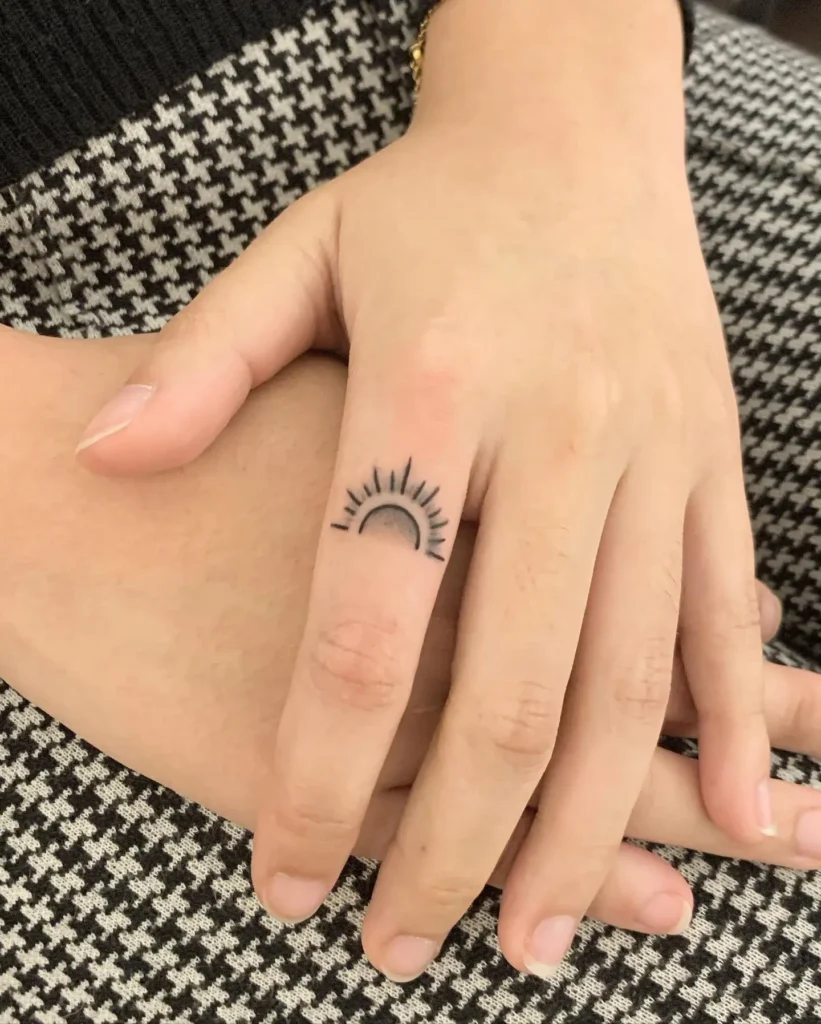 Close-up of a woman’s hand featuring a minimalist sun tattoo on the side of the ring finger. The black ink design shows a half sun with simple radiating lines, symbolizing warmth, energy, and new beginnings. Set against a stylish houndstooth outfit, the tattoo adds a subtle yet meaningful touch. This fine-line finger tattoo is ideal for women who love minimal, celestial-inspired body art with deep symbolism.


