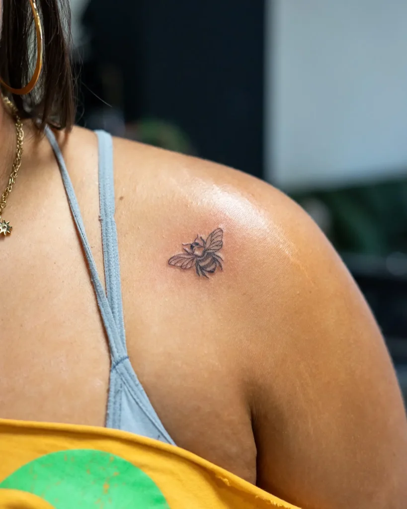 Small detailed bee tattoo on the shoulder of a woman, featuring delicate shading and fine linework.