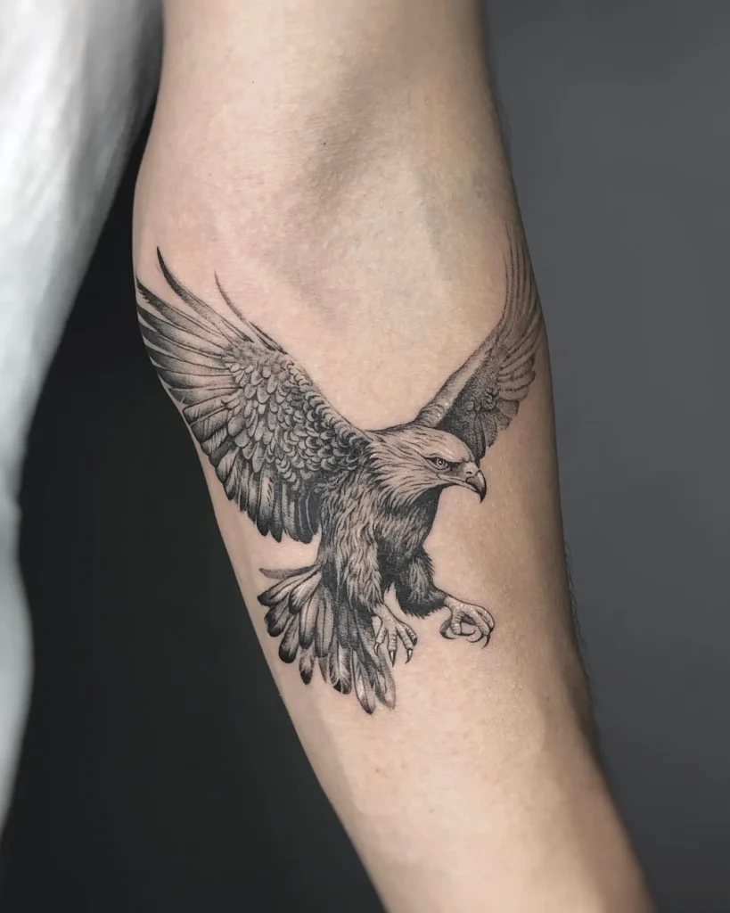 Realistic black and gray tattoo of a bald eagle in mid-flight, wings stretched wide and talons extended. Detailed shading highlights the texture of feathers and the fierce expression of the eagle. Placed on the upper arm with dynamic energy and bold symbolism of freedom and power.