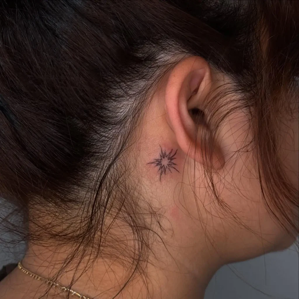 A small starburst tattoo behind a woman’s ear, featuring fine black lines that radiate in all directions, giving the look of a glowing light, with dark brown hair tied back for clear visibility.









