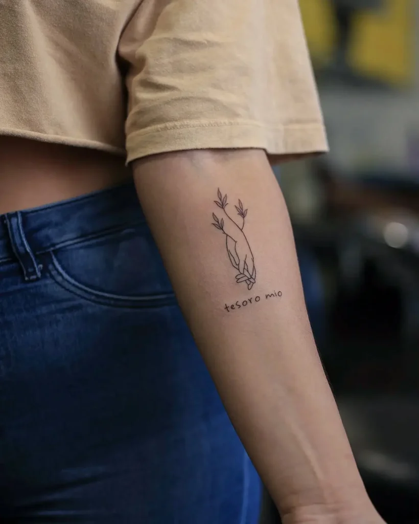Minimalist tattoo on the inner forearm shows two outlined hands gently clasped, with delicate leaves growing from the wrists. Below, “tesoro mio” is written in simple script. The design uses black ink and fine lines, making it a special option for mom and daughter tattoos unique, symbolizing affection, care, and treasured bonds.