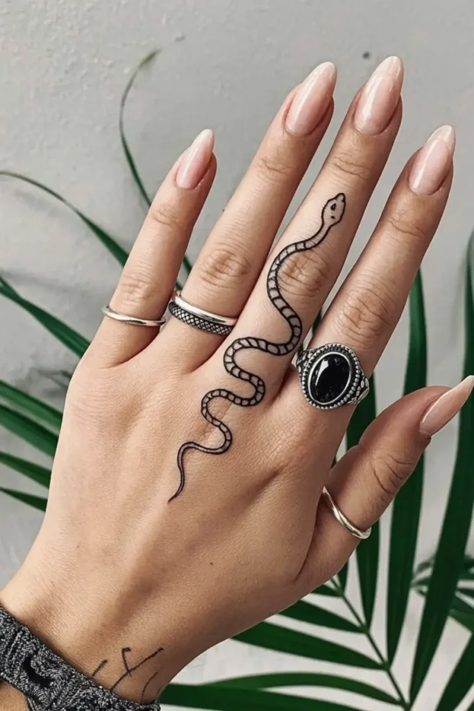 Close-up of a woman’s hand showcasing a bold black ink snake tattoo that curves from the middle finger down to the back of the hand. The fine-line design features a coiled serpent with minimal shading, symbolizing transformation and power. The woman wears silver rings and has almond-shaped nude nails, adding to the modern and edgy aesthetic. This striking finger tattoo idea blends symbolism with a fashion-forward vibe.









