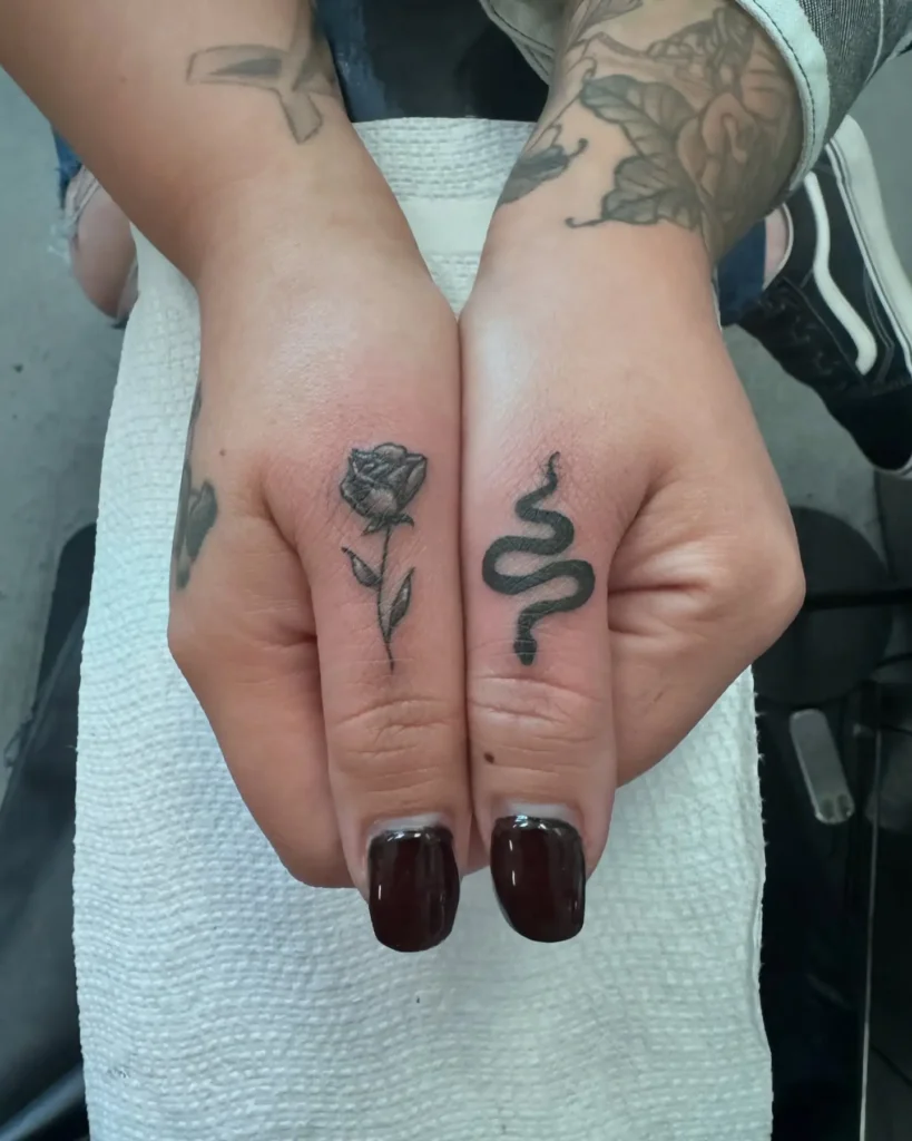 Close-up of two fingers featuring matching black ink tattoos—a detailed rose on the left index finger and a coiled snake on the right middle finger. Both tattoos use bold lines and soft shading for a striking yet balanced contrast. The woman has deep burgundy square-shaped nails, adding elegance to the edgy ink. These finger tattoo designs are ideal for women who love symbolic, contrasting themes like beauty and power.









