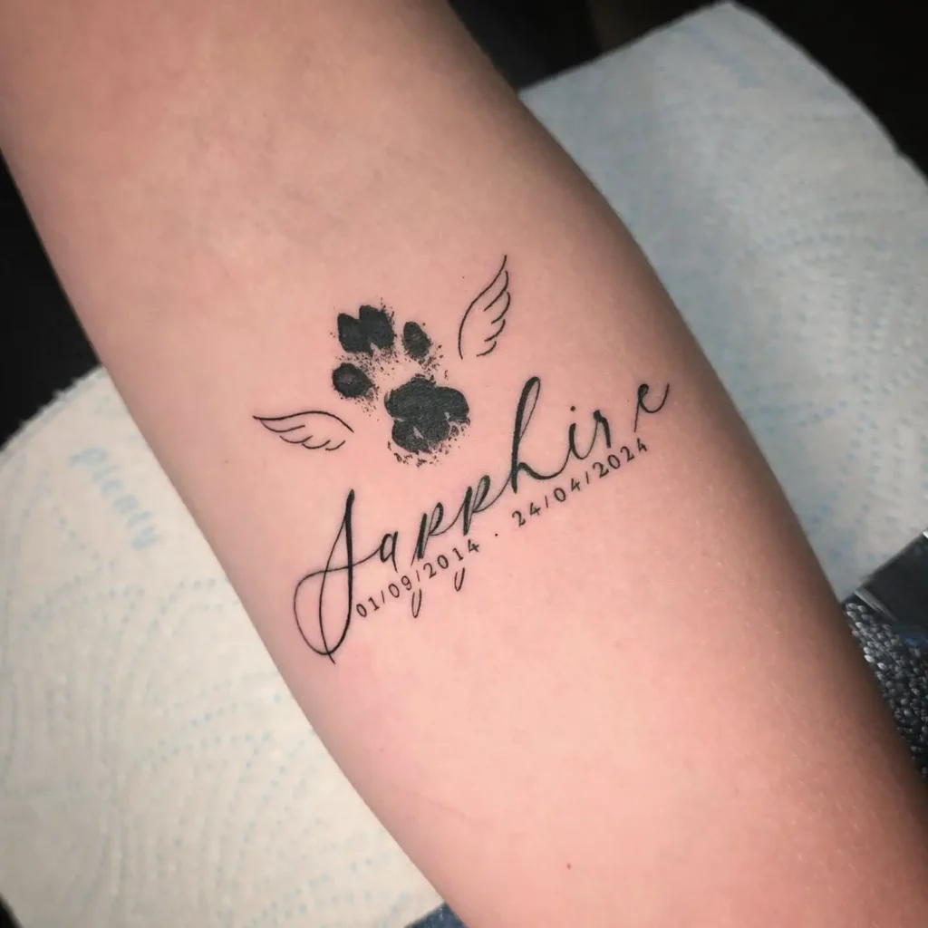 Tattoo of a black paw print with small wings above the name “Sapphire” and memorial dates, symbolizing remembrance in dog tattoo ideas memorial.