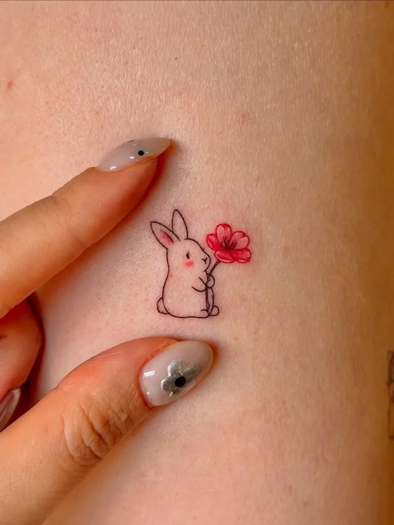 Minimalist micro tattoo of a cute white bunny holding a red flower, outlined in fine black ink with soft pink blush details. Delicate and tiny placement, perfect for a subtle and sweet aesthetic. Symbolizes innocence, love, and gentle energy.