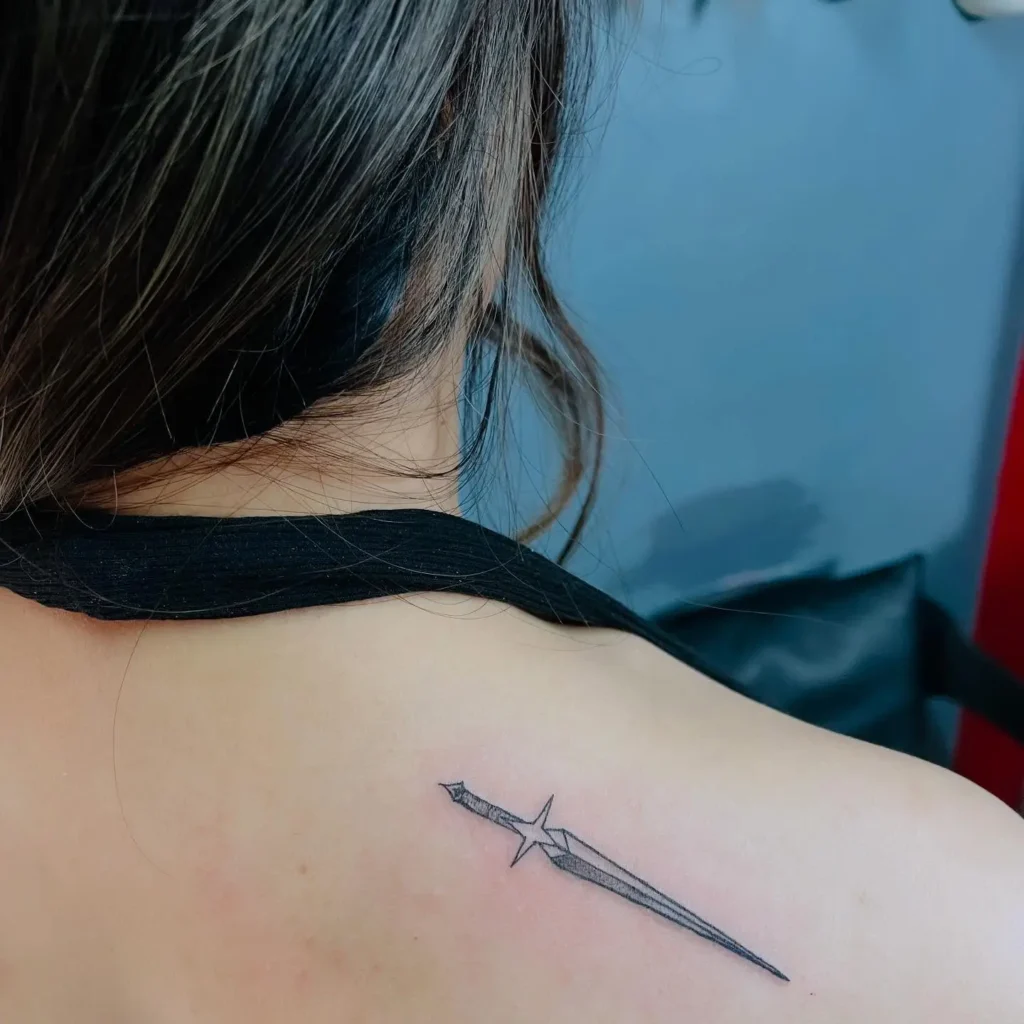 A minimalist sword tattoo on a woman’s upper back, positioned diagonally near her left shoulder blade. The black and gray ink design features clean, fine lines forming a sleek blade with a subtle starburst effect near the hilt, giving it a celestial and modern aesthetic. The woman is wearing a black halter top with her hair tied up, exposing the tattoo clearly against a soft-toned background.

