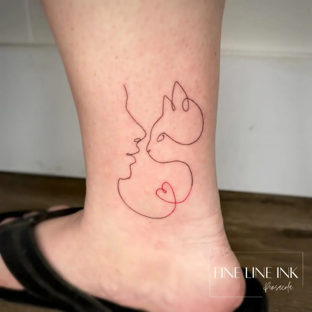Minimalist line tattoo on the ankle showing a woman’s face and a cat, connected by a small red heart.