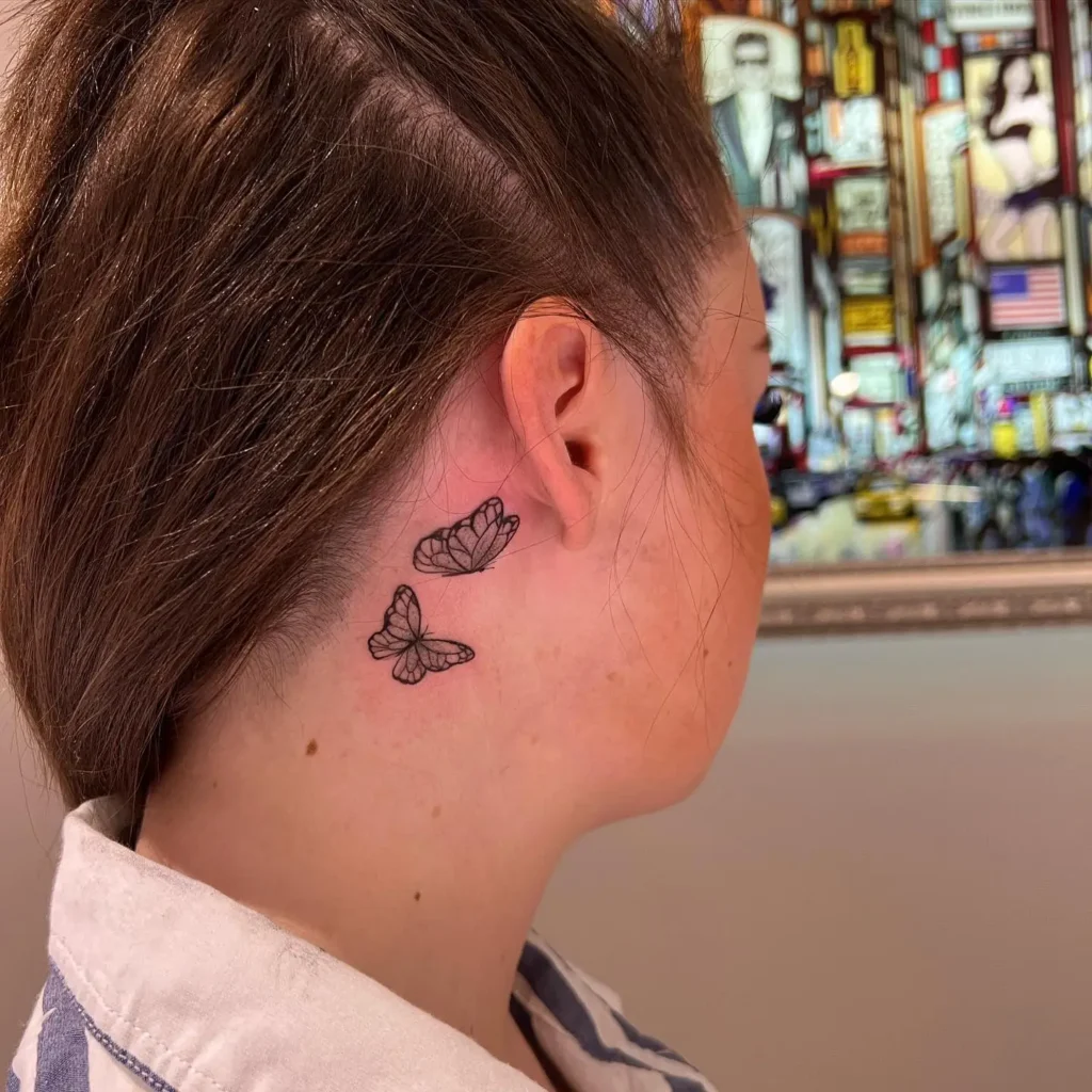 Minimal butterfly neck tattoo for women featuring two delicate linework butterflies below the ear, symbolizing freedom and softness.