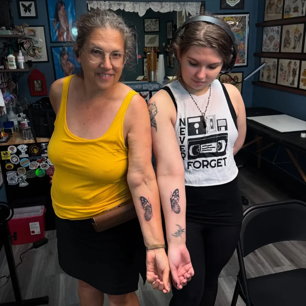 Mother and daughter showing off matching butterfly tattoos on their inner forearms, with realistic pink and black ink details. The tattoos symbolize connection and growth, highlighting a close relationship in a lively tattoo studio setting.