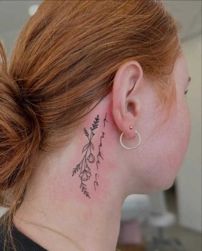 A vertical floral tattoo behind a woman’s ear, featuring fine line flowers and the handwritten phrase “i made it” in black ink, inked on fair skin with red undertones and auburn hair tied back.









