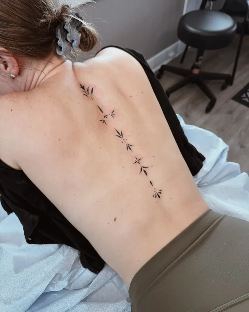 A minimalist botanical spine tattoo with small black leaves and star shapes running vertically down a woman’s back.