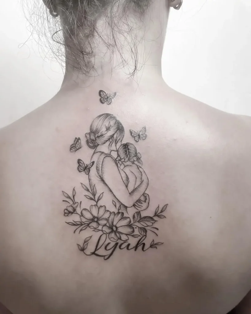 Back tattoo featuring a mother lovingly holding her daughter amid delicate flowers and flying butterflies. The name “Lyah” appears beneath the scene in script. Detailed black and grey artwork highlights the closeness of their bond. This design beautifully combines elements of mom and daughter tattoos butterfly, floral themes, and personal meaning.