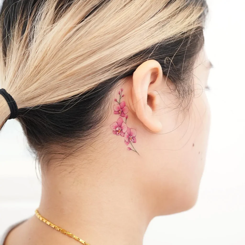 A watercolor-style pink flower tattoo placed behind a woman’s ear, featuring delicate orchid blossoms with soft shading and fine line stems, complementing her tied-back hair and golden necklace.









