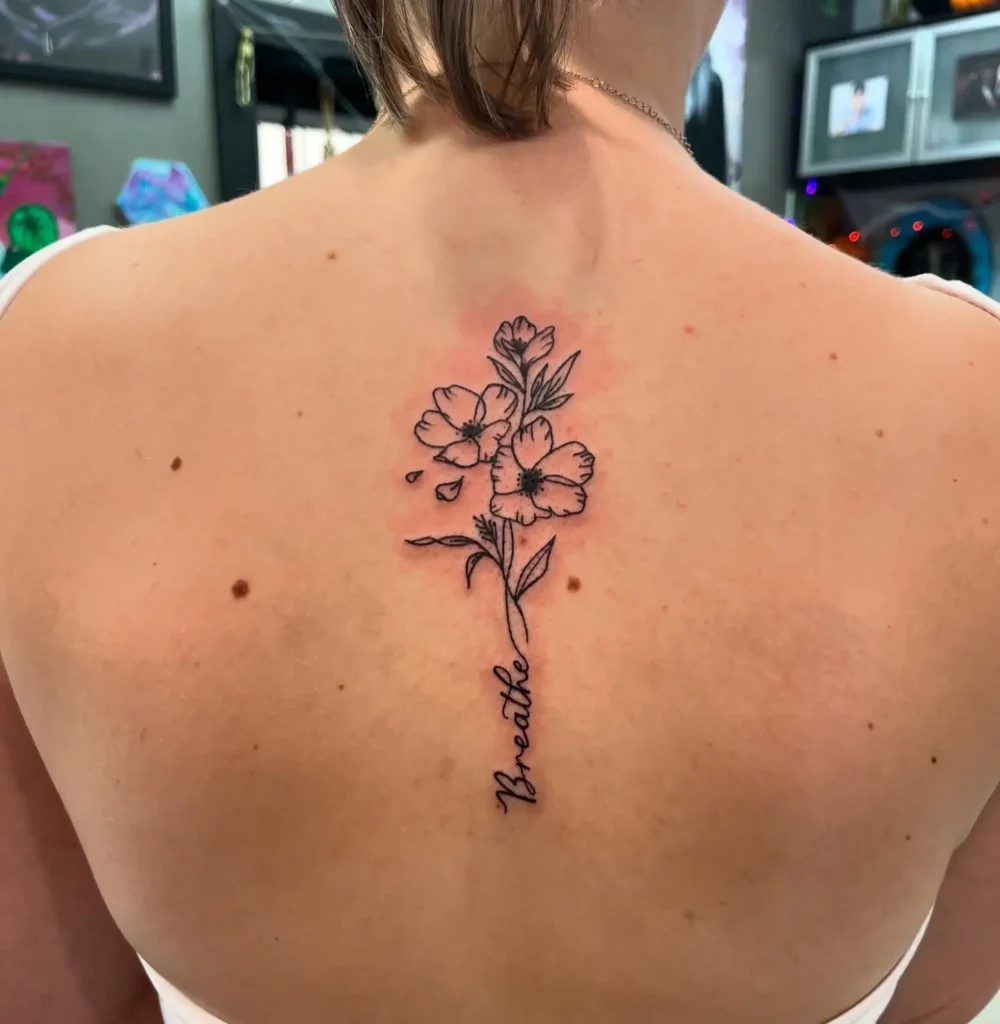 A spine tattoo featuring fine-line flowers and the word “Breathe” in script, running vertically down a woman’s upper back.