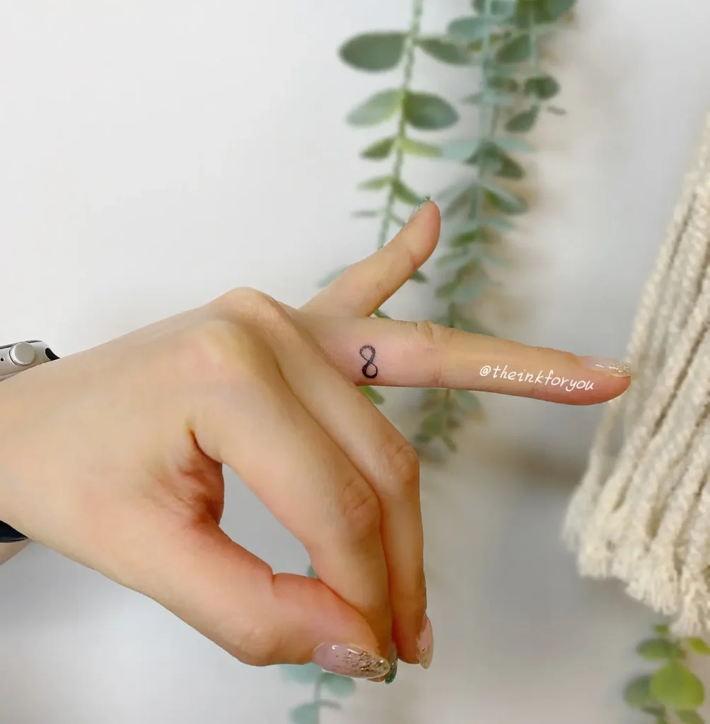 Close-up of a woman's hand showing a minimalist infinity symbol tattoo on the side of the middle finger. The small black ink design is simple yet meaningful, symbolizing eternity, balance, and endless love. Set against a soft background with hanging green leaves, the tattoo exudes elegance and subtlety. A perfect finger tattoo idea for women who prefer minimal, symbolic body art with personal significance.










