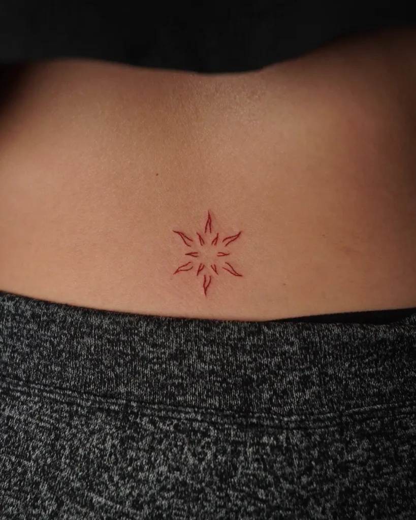 Tiny red sunburst tattoo inked on the lower back of a woman, featuring clean lines and a minimalist style.