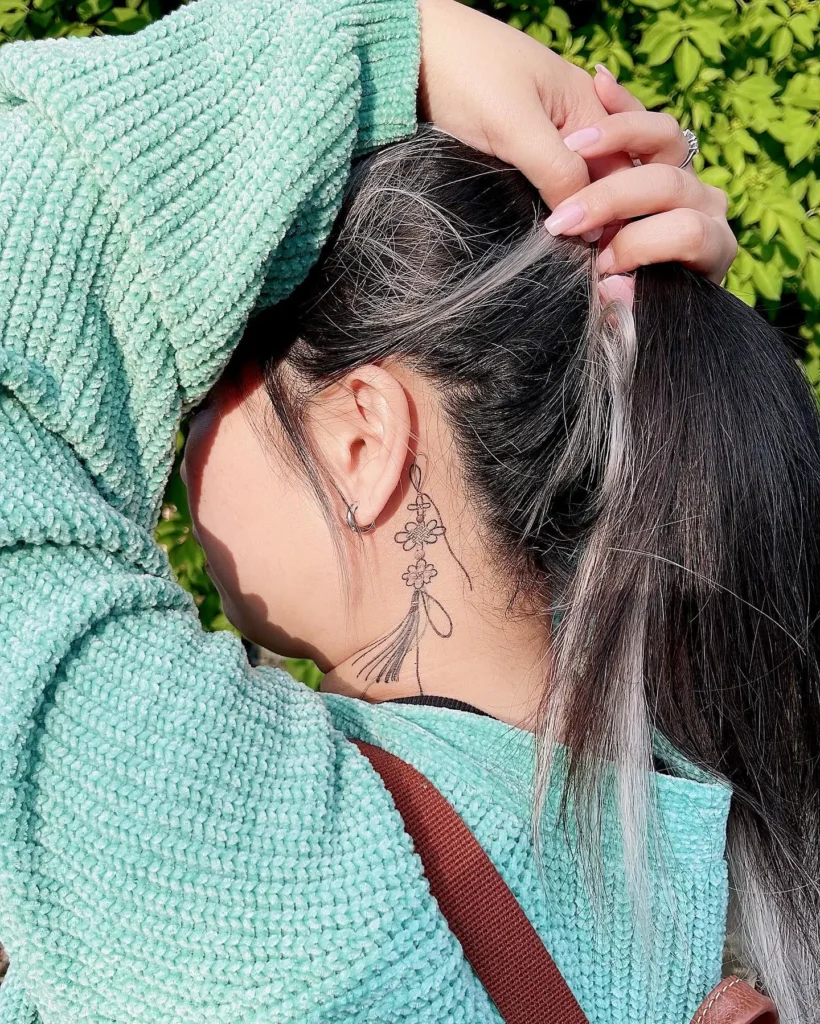 A fine line floral tattoo behind a woman’s ear, designed like a hanging ornament with three small flowers connected to a tassel, shown with her dark hair pulled up and a soft mint green sweater.









