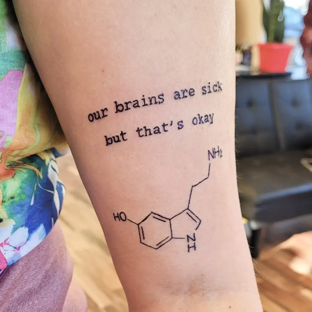 A tattoo with the words “our brains are sick but that’s okay” and a serotonin molecule diagram, symbolizing mental health awareness and acceptance.