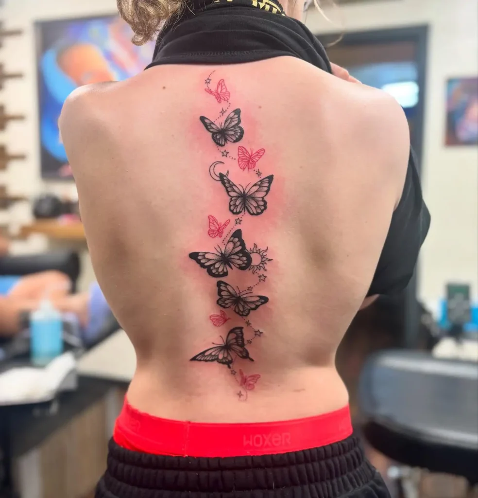 A cluster of black and red butterfly tattoos with stars and a crescent moon arranged vertically down a woman’s spine.