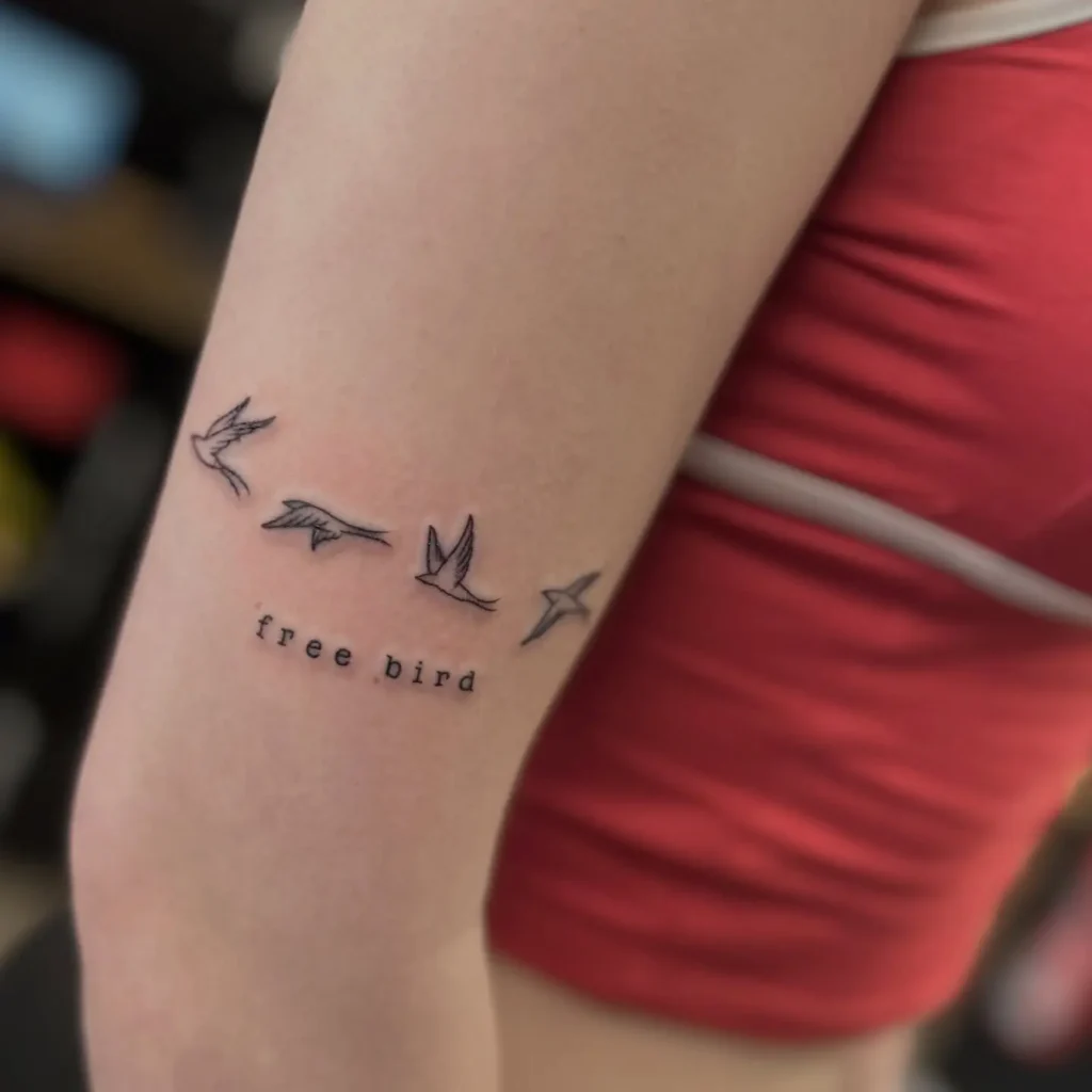 A tattoo on the upper arm with four flying birds above the phrase “free bird,” symbolizing freedom, independence, and self-liberation.