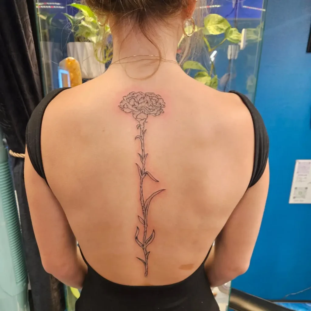 Vertical carnation flower tattoo running along the spine of a woman’s back, showing detailed petals and leaves.