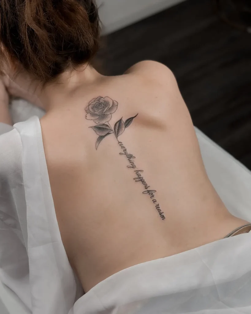 A spine tattoo featuring a shaded rose at the top and a script quote forming the stem, running down a woman’s back.