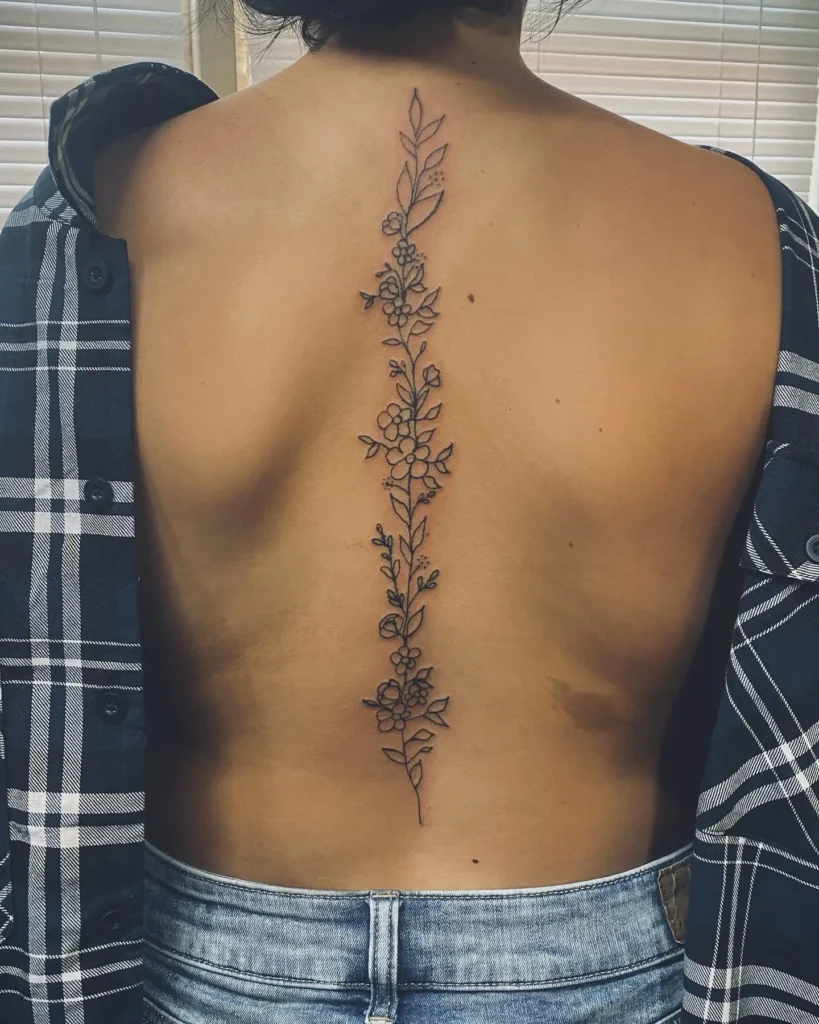 A fine-line spine tattoo with a vertical vine of leaves and small flowers running down a woman’s back.