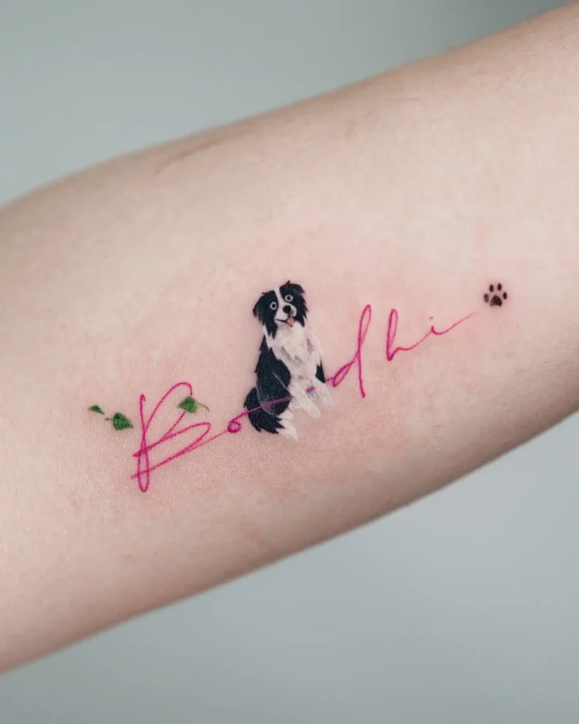 Minimalist tattoo of a small black and white dog above pink script spelling “Bodhi,” with green leaves and a tiny paw print on light skin.