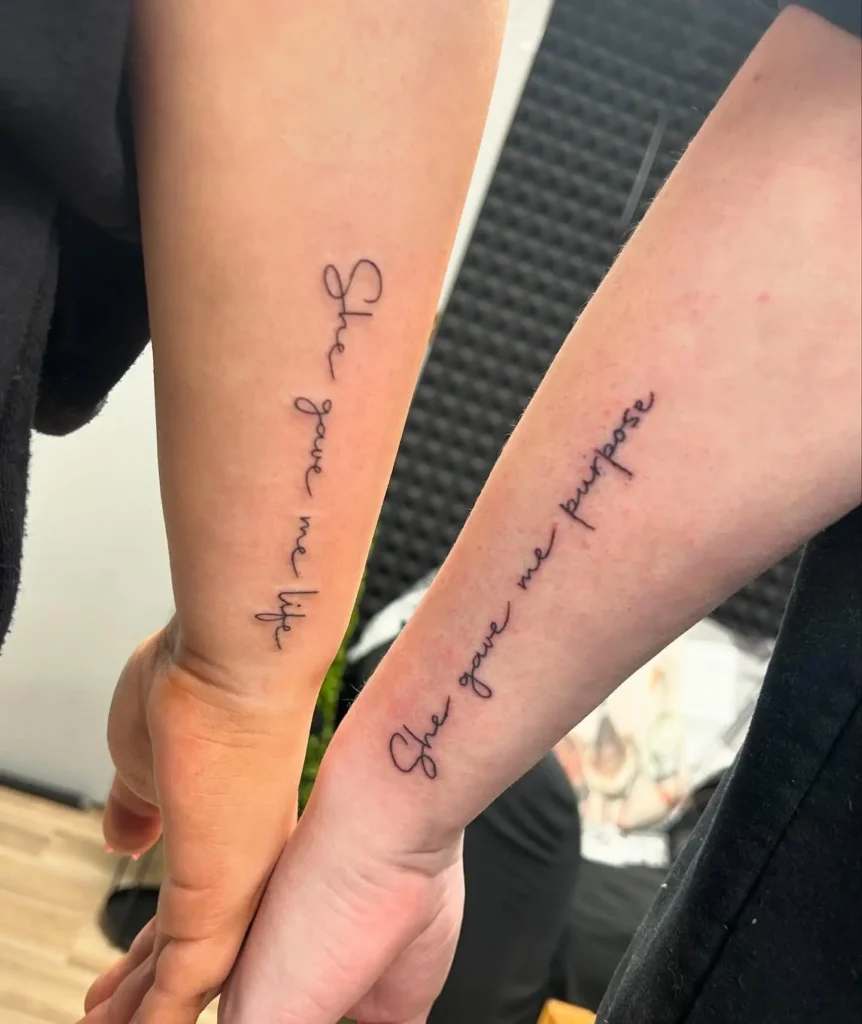 Two forearms with matching tattoos written in elegant cursive. One arm reads “She gave me life,” while the other says “She gave me purpose.” The design uses black ink and simple lines, making it meaningful and easy to read. This tattoo style is a heartfelt choice for mom and daughter tattoos, focusing on their unique bond through words.