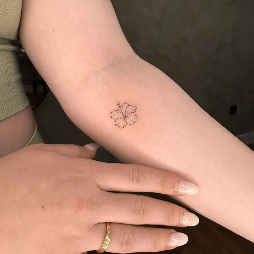 Minimalist black outline hibiscus flower tattoo placed on the inner arm of a woman.