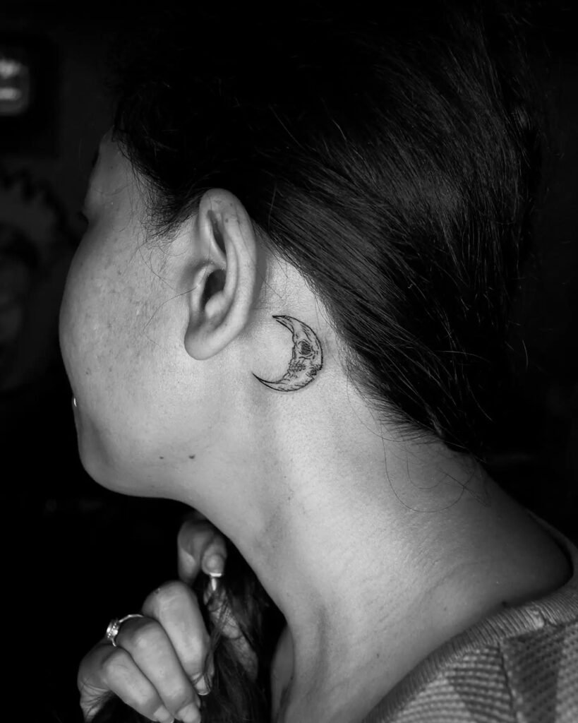 A finely detailed crescent moon tattoo placed behind a woman’s ear, shown in black ink with textured shading, captured in a black-and-white photo emphasizing the lunar design and skin contrast.