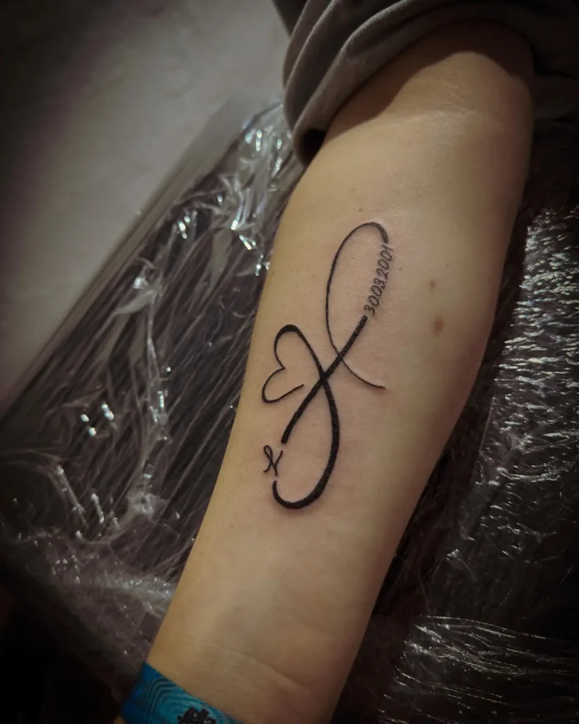 Black ink infinity symbol tattoo with a heart and a name, placed on a woman’s inner forearm.