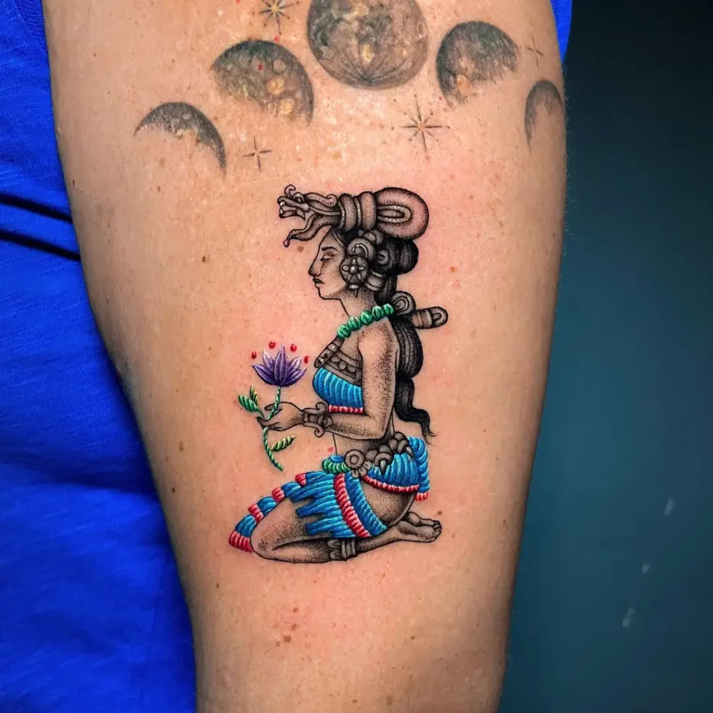 Colorful tattoo of Mayan goddess Ixchel kneeling with a flower, featuring embroidery-style textures and traditional Mexican elements on upper arm.
