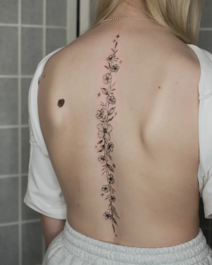 A vertical wildflower spine tattoo with fine-line blooms and leaves on a woman’s back.