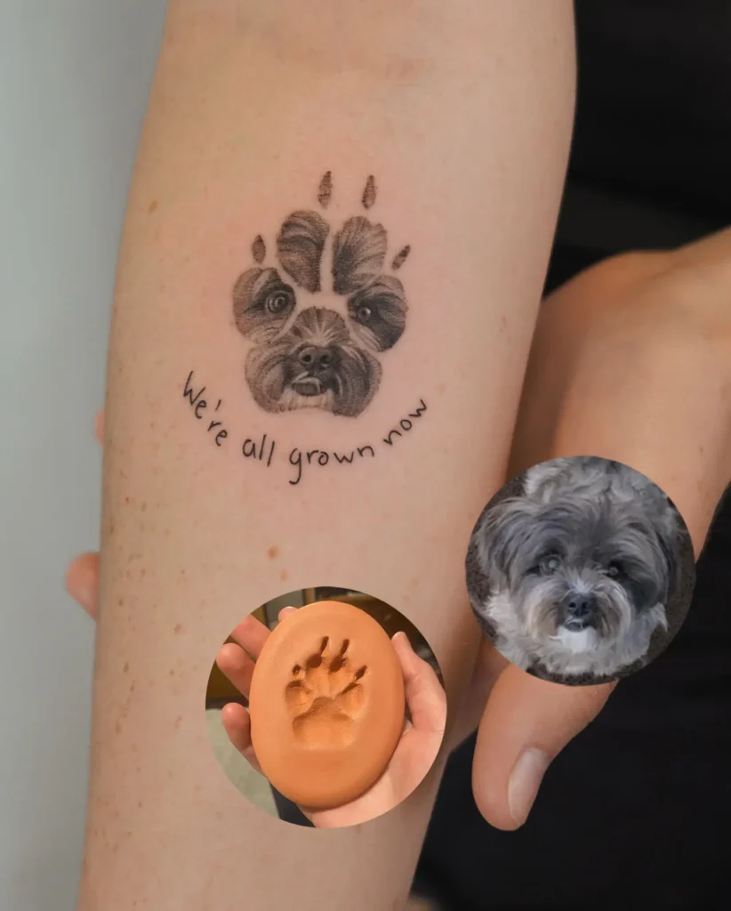 Tattoo of a realistic dog’s face blended into a paw print, with the phrase “We’re all grown now” beneath, on light skin, symbolizing lasting memories and love for a pet.
