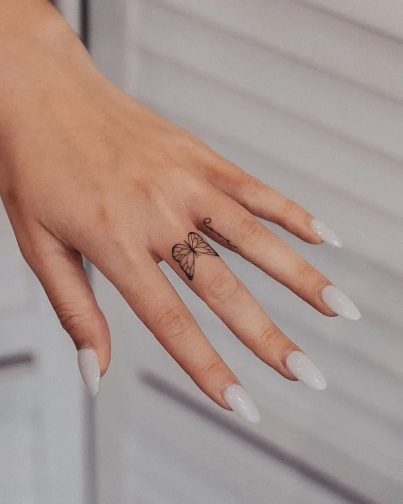 Close-up of a woman's hand showcasing a delicate butterfly finger tattoo on the middle finger. The tattoo features fine black ink linework, creating an elegant and minimalist look. The woman has long almond-shaped nails painted in glossy white, enhancing the overall soft and feminine aesthetic. Ideal inspiration for those seeking finger tattoos for women with butterfly elegance and subtle detail.








