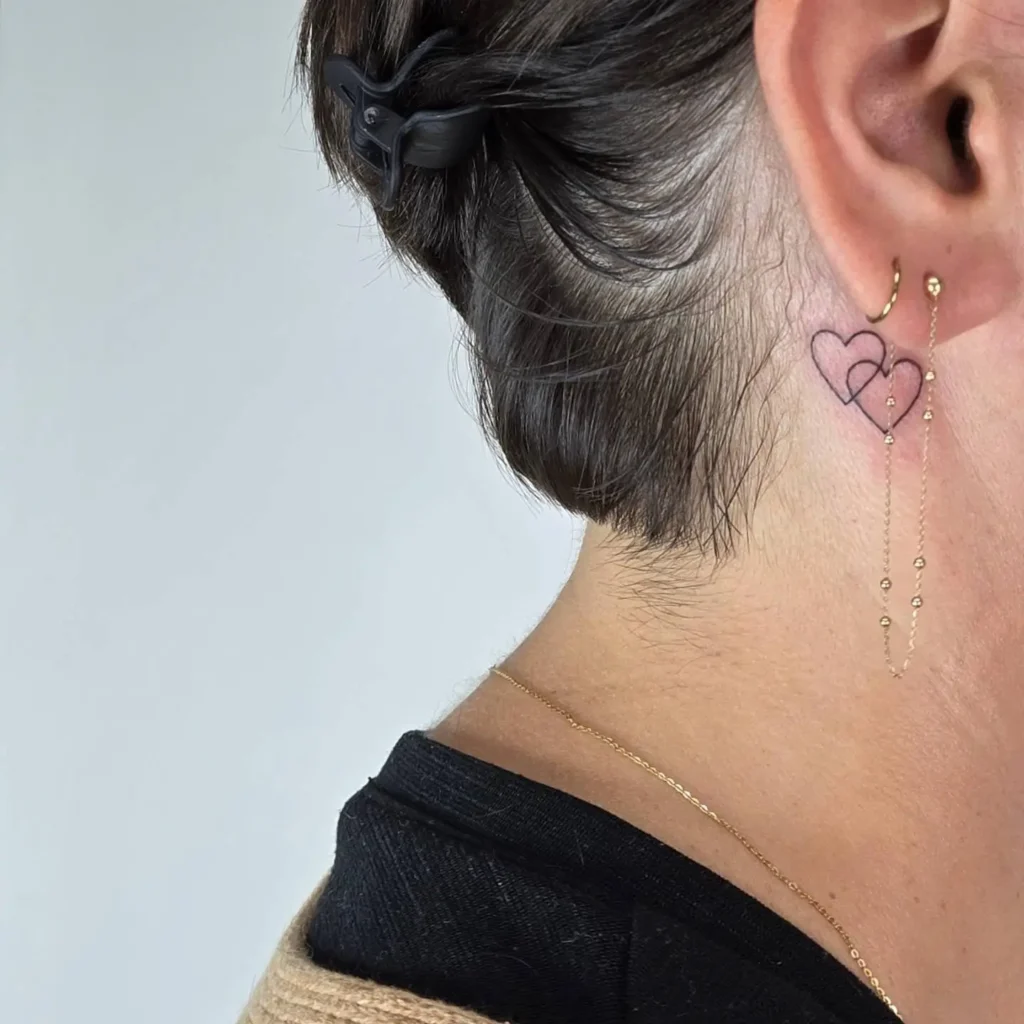 A behind the ear tattoo of two interlocking hearts outlined in pink and black ink on a woman’s neck, paired with gold chain earrings and a short hairstyle clipped back for clear visibility.









