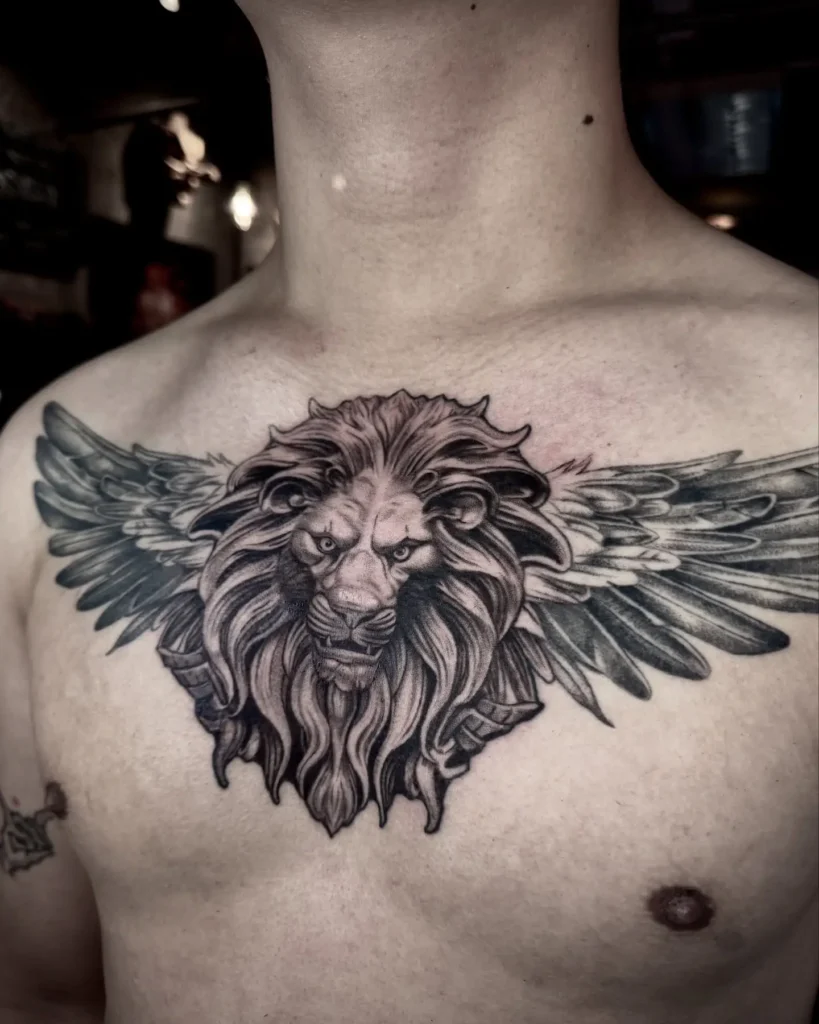 Chest tattoo for men showing a hyper-detailed lion head with large outstretched wings; intense shading in the mane and feathers emphasizes power, freedom, and leadership in a symmetrical black-and-grey design.