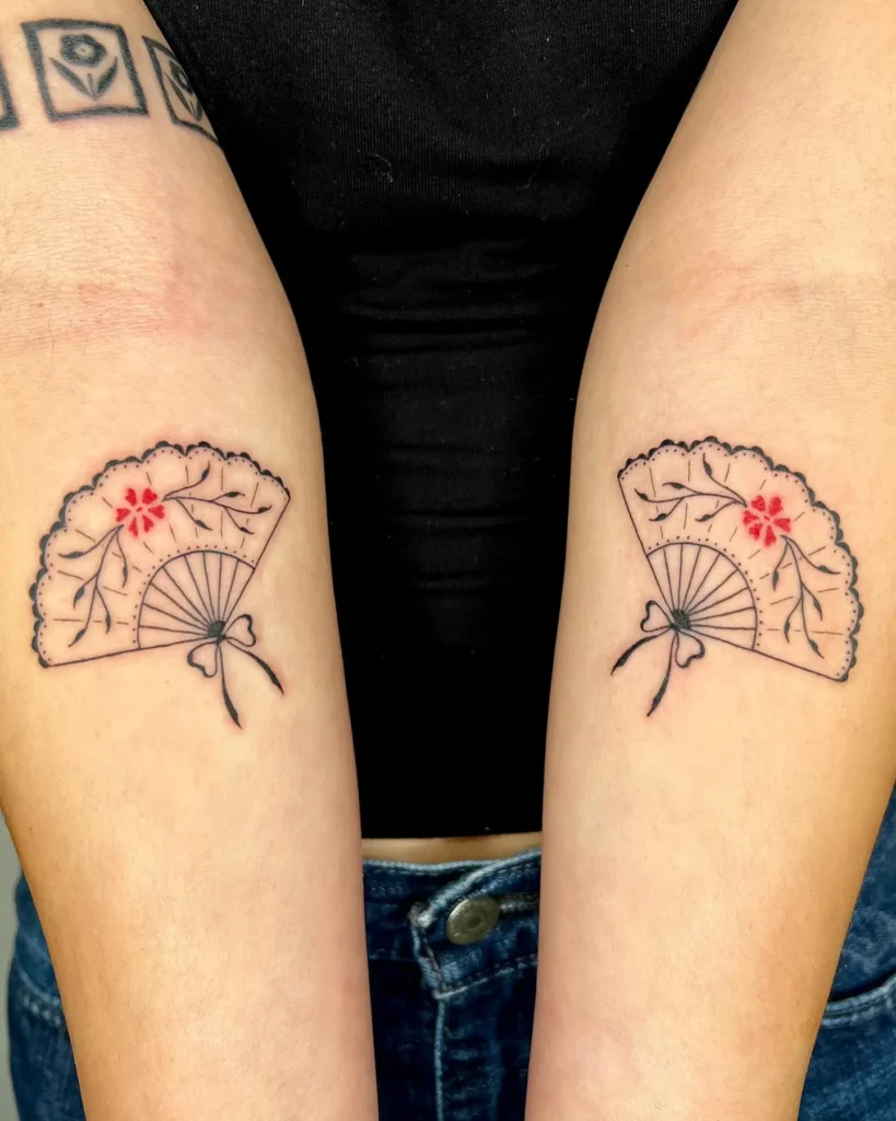 Matching tattoos on two forearms featuring an open hand fan outlined in black, detailed with a small red flower and leafy accents. Each fan is tied with a thin ribbon at the handle. This design blends tradition with elegance, making it a special choice for mom and daughter tattoos that express unity, culture, and personal style.