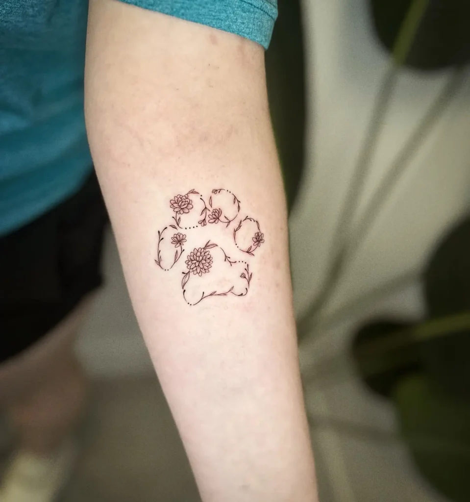 Fine line tattoo of a dog’s paw print with detailed floral accents and vines, placed on the forearm, representing love for dogs and nature.