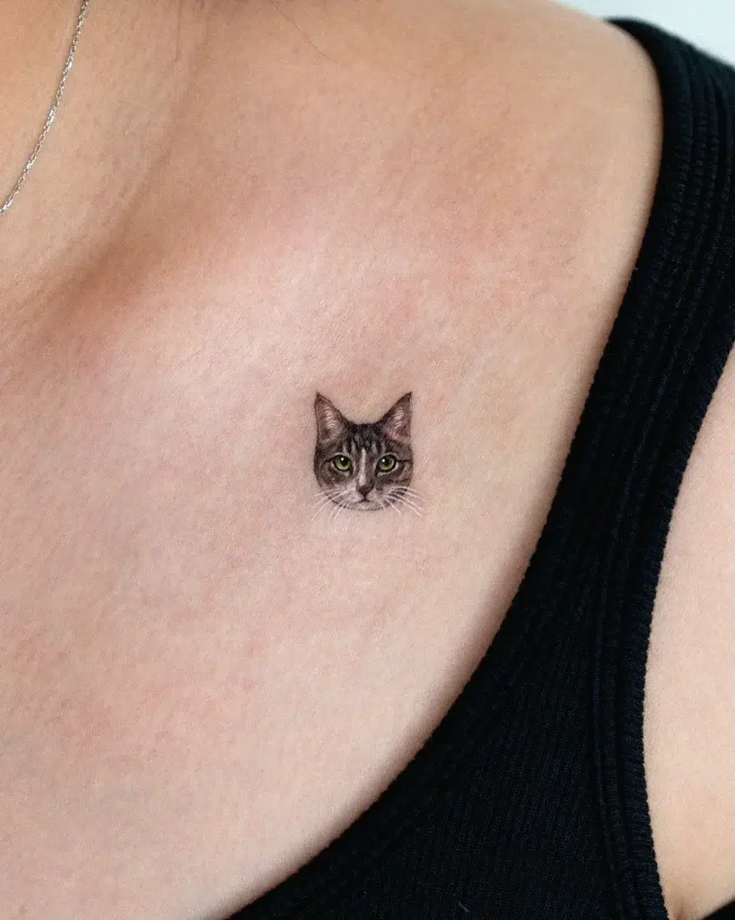 Small realistic cat face tattoo placed near the collarbone. Detailed black and grey fur with soft green eyes. Minimal and meaningful—great for animal tattoo ideas simple and personal. Perfect size for upper chest or discreet body art honoring pets or favorite animals.








