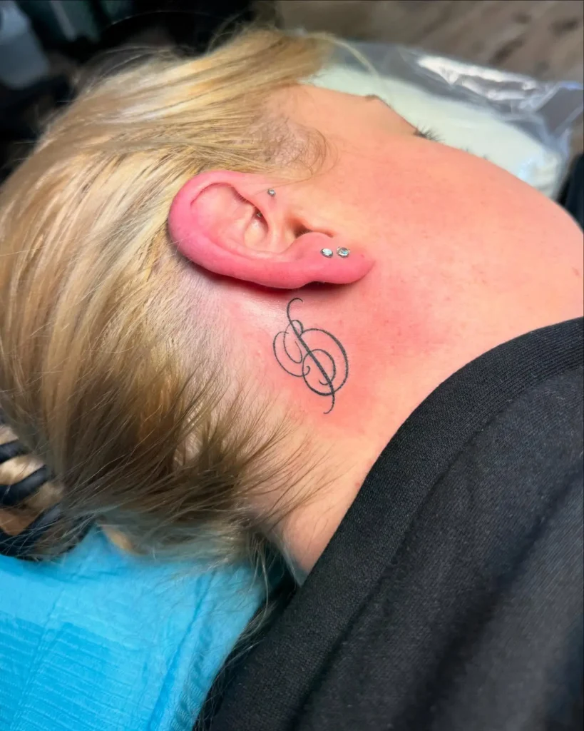 A behind the ear tattoo featuring a black treble clef with cursive initials, inked on a woman with blonde hair lying on a tattoo chair, showcasing clean linework and subtle personal flair.









