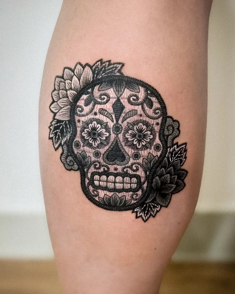 Black embroidered sugar skull tattoo with floral patterns and bold petal accents, inspired by Mexican tattoo ideas and Day of the Dead traditions.